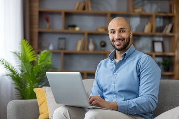 Smiling young man working or studying from his home office, relaxed on a sofa with a laptop, typing and engaging in remote work, online learning or video calls in a modern living room