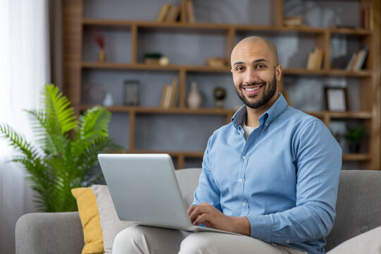 Smiling young man working or studying from his home office, relaxed on a sofa with a laptop, typing and engaging in remote work, online learning or video calls in a modern living room - Powered by Adobe