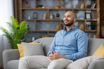 Happy bald man with a beard relaxing on a comfortable sofa in a modern living room, smiling and...