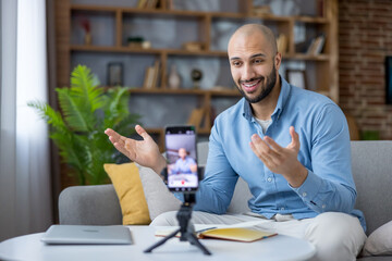 Young bald bearded man smiling and speaking to camera while recording a live stream or online webinar with smartphone on tripod in a casual home living room setting