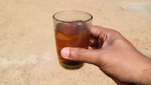 Close up of hand holding glass with brown contaminated undrinkable dirty drinking water on a hot summer dry ground soil background. Water crisis, scarcity, shortage, drought and less rainfall concept.