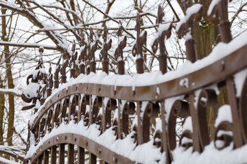 An exquisite wrought-iron fence with decorative elements is covered with a thick layer of fresh snow against a background of winter trees. 