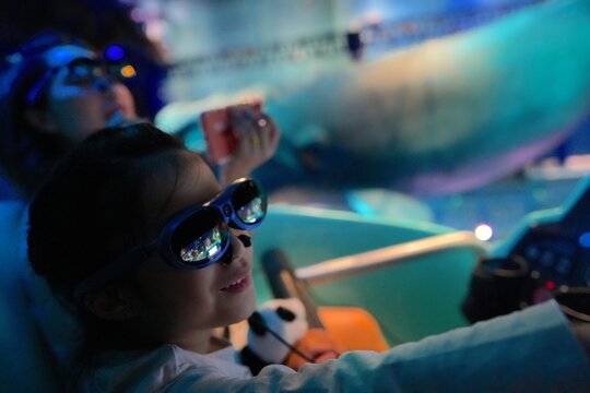A young child wearing 3D glasses enjoying an immersive cinematic or theme park ride experience, illuminated by colorful lights in a futuristic indoor environment with wonder entertainment in cinema.