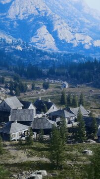 panoramic mountain settlement with winding road rows of stone houses descending valley beneath towering peaks, sparse trees and travel route slicing scene,