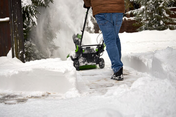 Man clearing a narrow driveway with an electric snow blower after heavy snowfall. Winter maintenance in a residential area, deep snowbanks, flying snow spray, cold weather and daily life routine