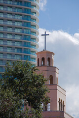 Obraz premium Chapel Cross Beside Modern High Rise Building Against Bright Blue Sky in St. Petersburg, FL. Peach-colored church tower with a cross sits beside a curved glass high-rise, under a clear blue sky. The s
