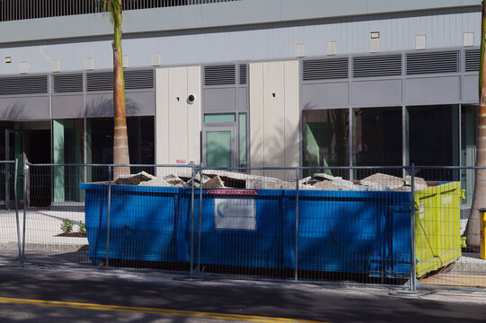 Urban Construction Dumpster Behind Modern Building With Fenced Site On City Street. Blue and yellow dumpsters filled with gravel sit behind a metal fence in front of a modern building. A construction 