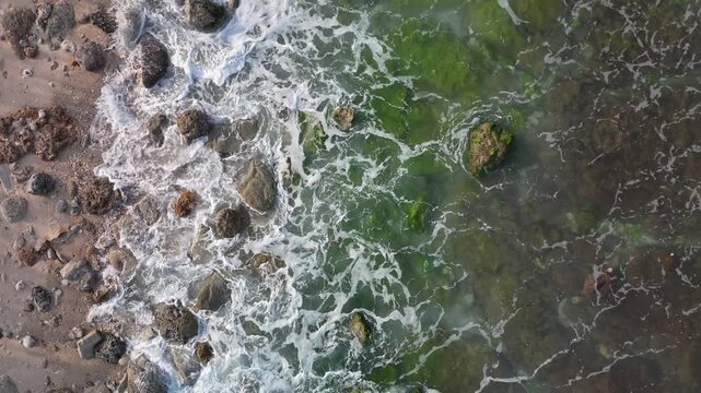 Experience the stunning top down aerial view of ocean waves crashing against mossy rocks and a pebble beach showcasing the rugged beauty of the coastal shoreline