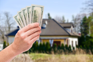 Hand holding US dollar banknotes in front of a residential house.