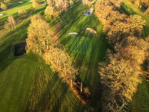 Aerial drone view of golf course green at golden hour with long tree shadows and pond