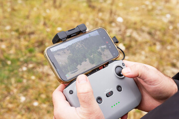 Person's hands holding a drone remote controller with a smartphone attached against a forest backdrop © Elena