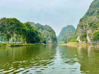 Obraz premium Peaceful Ngo Dong River leading the way to lush green mountains in Tam Coc Ninh Binh. The photo is taken from a central view of the river, where green trees on the banks reflect in the water