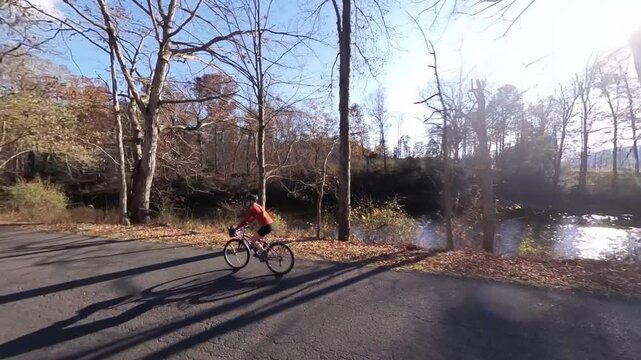 A mature woman bikes on a road by a river in West Virginia surrounded by trees in autumn with clear skies and warm sunlight.