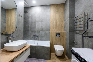 Modern bathroom with grey tiles, wooden accents, white sink, bathtub, wall-mounted toilet, heated towel rack, round mirror, and built-in washing machine &mdash; minimalist design