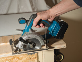 Hand Using Cordless Circular Saw to Cut Plywood; Close-up of a worker's hand holding and operating a battery-powered circular saw to cut a wooden plank in a home workshop or construction site.