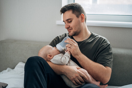 A father feeds his newborn son from a bottle of milk while sitting on the bed. The concept of caring for infants by fathers