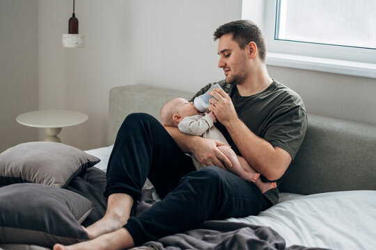A father feeds his newborn son from a bottle of milk while sitting on the bed. The concept of caring for infants by fathers