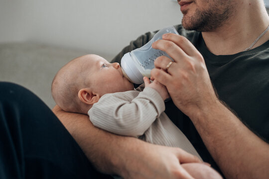 A father feeds his newborn son from a bottle of milk while sitting on the bed. The concept of caring for infants by fathers