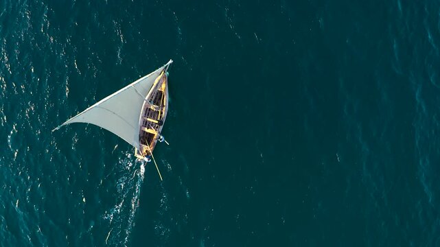 Aerial view of a traditional dhow with its white sail cutting through the deep blue ocean waters under the golden sunlight, Nampula, Nampula Province, Mozambique.