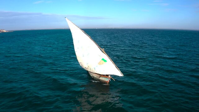 Aerial view of a dhow with a white sail cutting through the deep blue sea under a clear sky, creating a striking contrast, Nampula, Nampula Province, Mozambique.