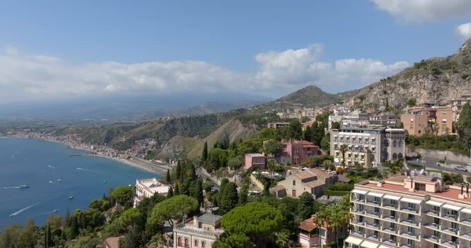 Panorama of the town of Taormina in Sicily, with a view of the Ionian Sea. Elegant buildings and hotels are nestled among lush Mediterranean trees on the hillside, overlooking a long coastline.