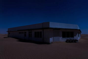 Closed shop storefront standing alone in a desert at night, surrounded by sand dunes under a deep blue, moody sky.