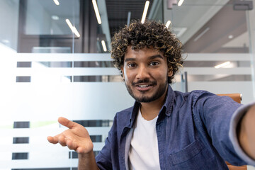 Close-up photo of a young smiling Indian man in an office, talking to a camera he is holding in his hand