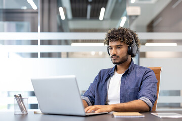 Concentrated young Indian man wearing headphones working on laptop in office
