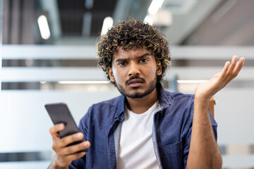 Close-up photo of a young Indian man holding a phone in his hand, looking at the camera with dissatisfaction and disappointment