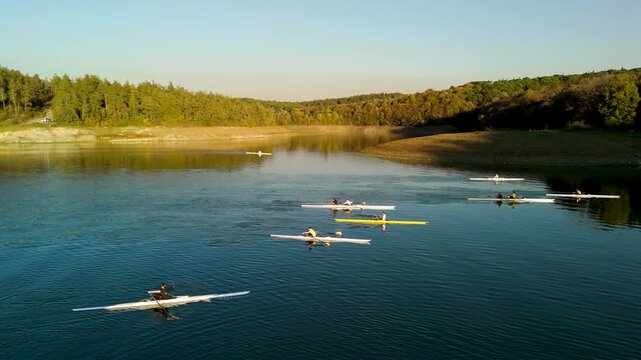 High angle view of colorful rowing shells practicing on a calm lake with sunlit forested hills in the background.