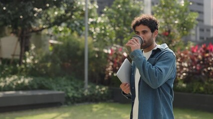 Portrait of happy Hispanic man with digital tablet drinks coffee in green city park. Latin male student holds mobile device as walks along spring downtown street slow motion - Powered by Adobe
