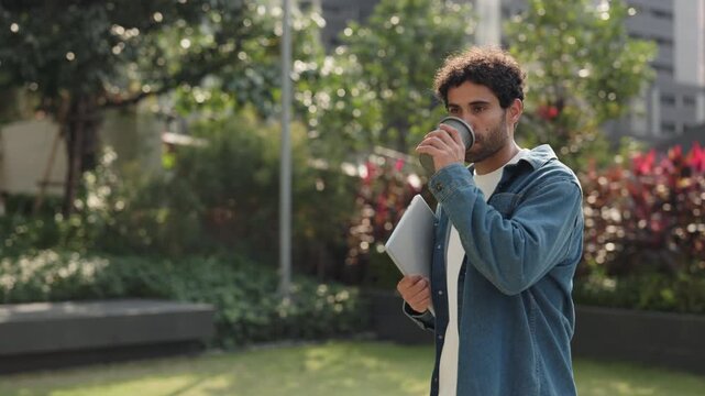 Portrait of happy Hispanic man with digital tablet drinks coffee in green city park. Latin male student holds mobile device as walks along spring downtown street slow motion