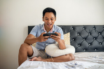 Asian young man sits on a bed, smiling and happy while using a smartphone with wired earphones for mobile gaming.