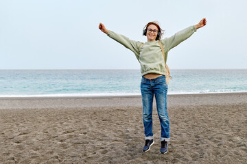 Smiling middle-aged female in headphones listening music and dancing while walking near the sea.