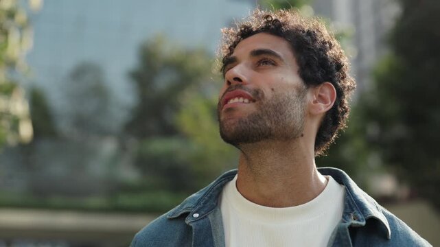 Portrait of happy Hispanic man in casual attire looks upward in city park. Smiling Latin American guy with beard gazes at skyscrapers in summer urban district. Travel destination