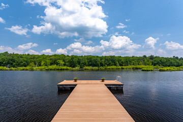 Wooden dock extending into quiet lake with forest shoreline and dramatic clouds, peaceful summer landscape with blue sky and calm water