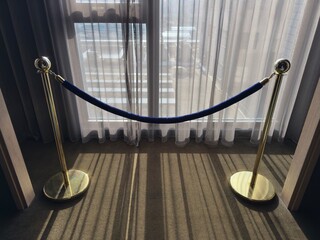 VIP Velvet Rope and Sheer Curtains: Navy blue velvet rope barrier with gold posts in front of sheer curtains and bright window light.