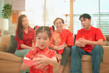 Young girl opens a red envelope gift while her family chats happily together on background
