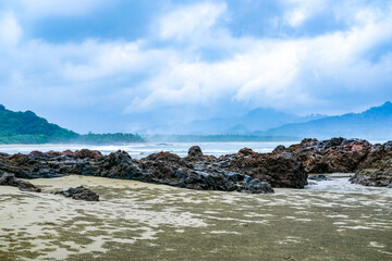Rocky shoreline at Rajegwesi Beach inside Meru Betiri National Park, featuring coral rocks, calm waves, tropical forest backdrop, and overcast skies along the southern coast of Java.