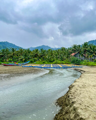 Serene fishing village scene on South Java coast under cloudy skies, shallow river mouth flowing into sandy beach, palm trees lining the shore, distant blue boats, and misty mountains. Rajegwesi Beach