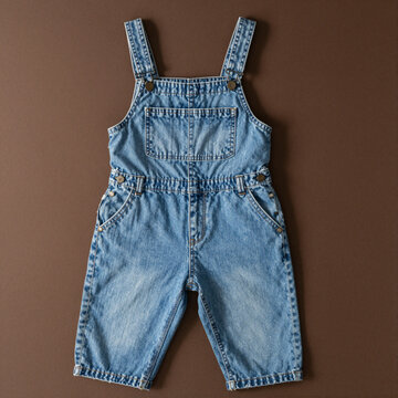 Light-wash denim romper with adjustable buttoned straps, chest pocket and belt loops laid flat on a brown backdrop