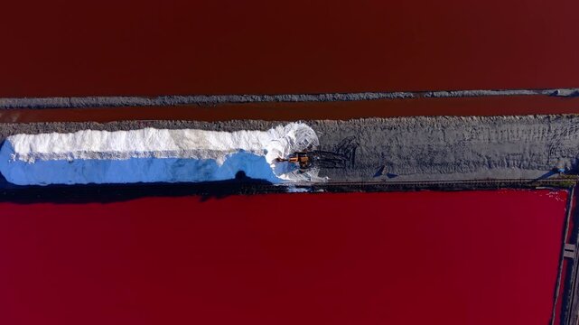 Aerial veiw of a yellow excavator on a narrow vertical path. The machine is flanked by deep red water and white salt.