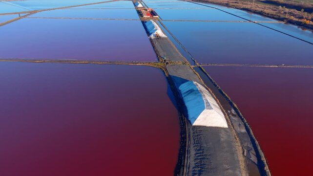 A wide top-down aerial view of rectangular salt basins. The water is a deep pink due to microorganisms with the white pyramids of harvested salt.