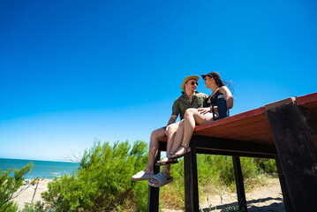 A man and a woman are sitting on a wooden platform by the beach
