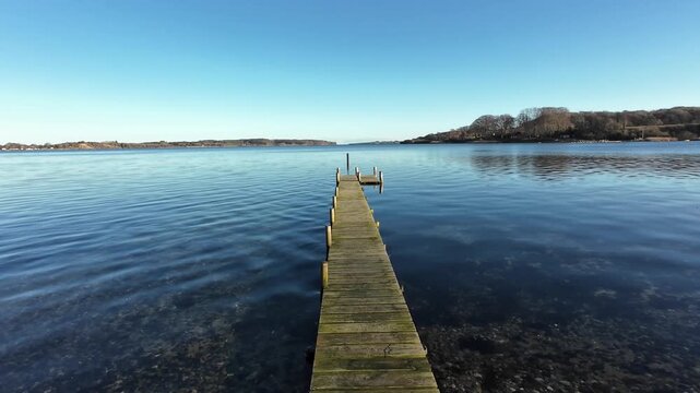rone video of wooden pier on clear lake surrounded by forest and blue sky in Denmark