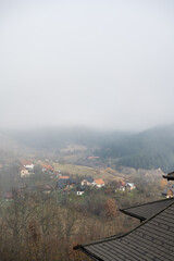 A serene rural mountain vista featuring small homes nestled amid grassy clearings and bare trees, with a dense pine forest climbing a steep ridge shrouded in morning mist. 