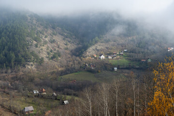 A misty mountain valley scene with scattered rural houses perched on a hillside and evergreen forest rising into low fog above. Autumn trees and green fields contrast with rocky slopes.