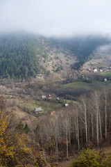 Fog rolling over a wooded mountain slope with patches of green pasture and a handful of quaint village houses below. 
