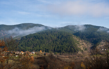 A misty mountain valley scene with scattered rural houses perched on a hillside and evergreen forest rising into low fog above. Autumn trees and green fields contrast with rocky slopes.