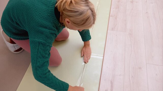Young woman kneeling while taping the seams of thermal insulation underlay panels with clear adhesive tape before installing new laminate flooring during a home renovation project